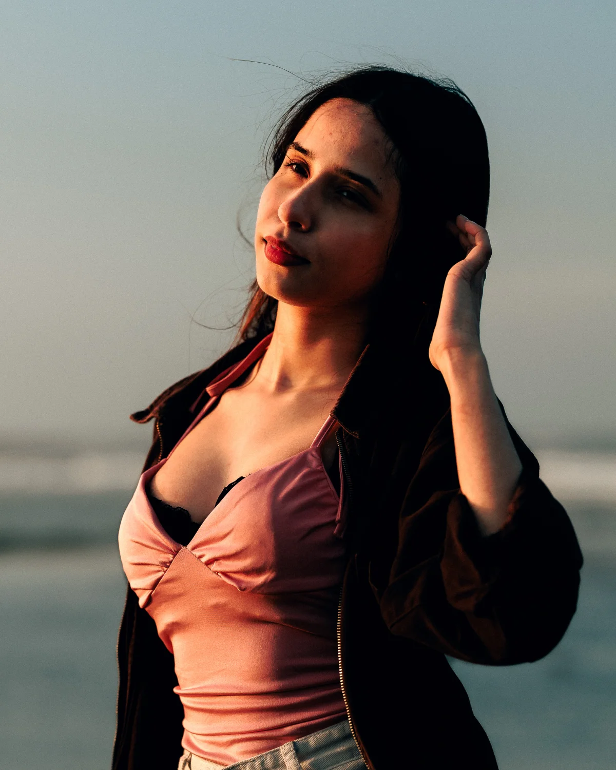 A woman with dark hair in a pink satin top and dark jacket posing outdoors during golden hour.