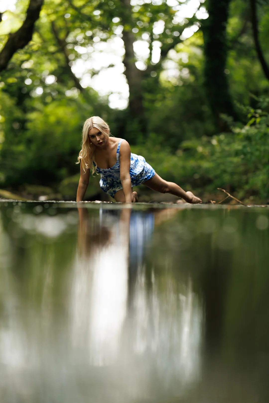 Blonde woman in a blue patterned dress posing by a forest stream with a reflection in the water.