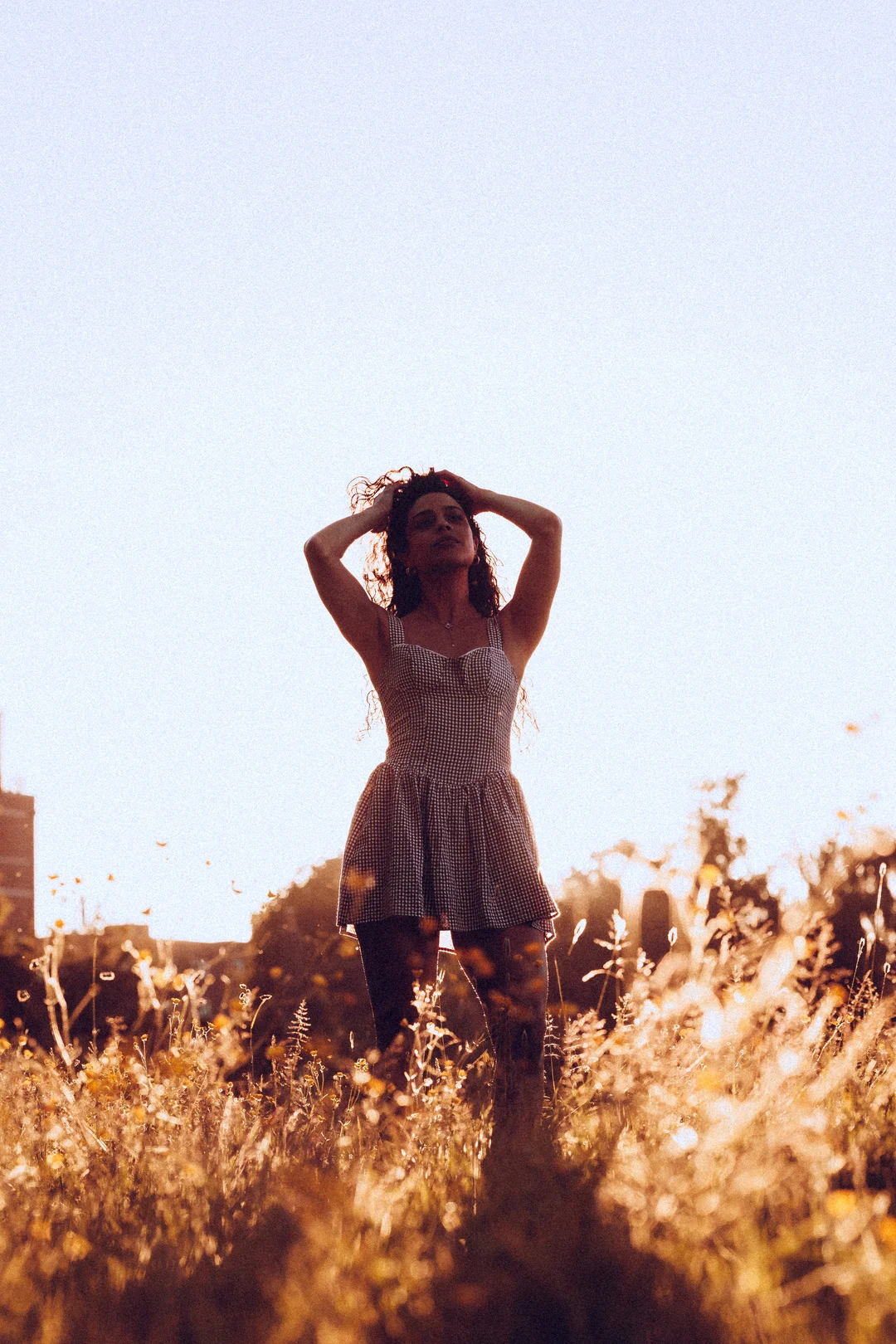 A woman in a checkered dress posing in a golden field during sunset, hands in her hair, backlit by soft light.