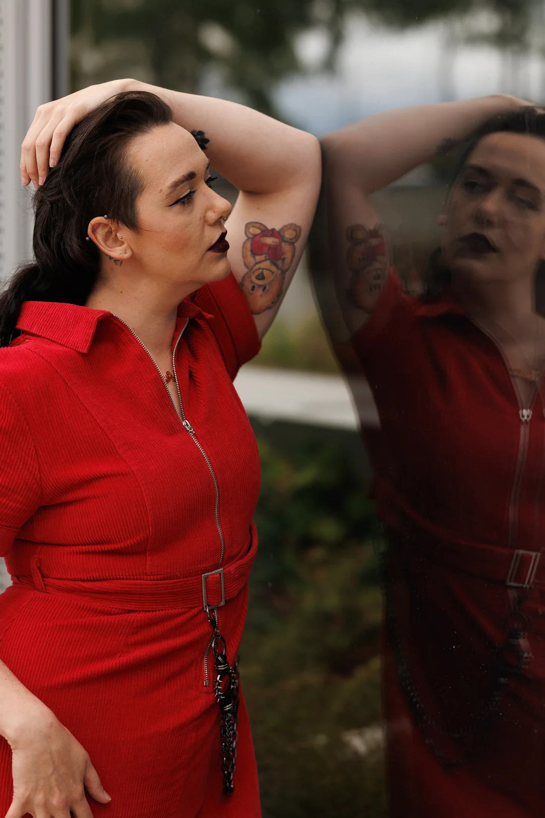 A person in a red corduroy jumpsuit posing against a reflective glass window, showcasing their profile and a tattoo.
