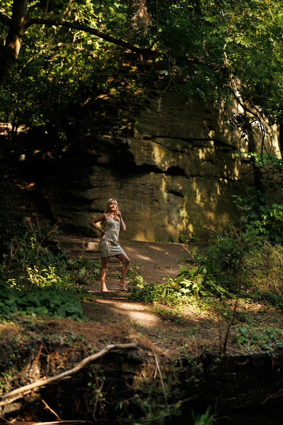 A woman in a short patterned dress posing in a sun-dappled forest in front of a rock wall.
