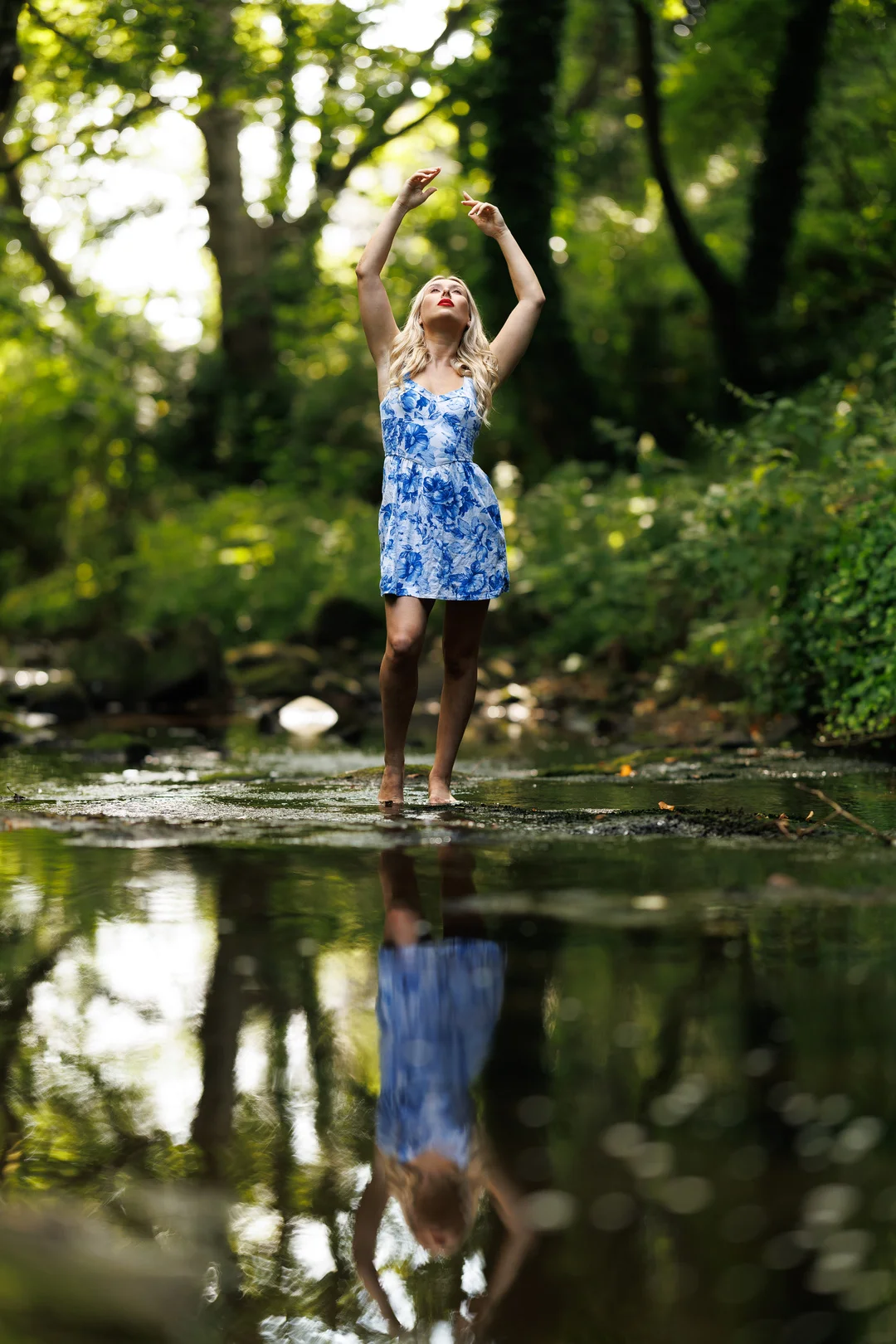 Blonde woman in a blue floral dress posing in a forest stream with a clear reflection in the water.