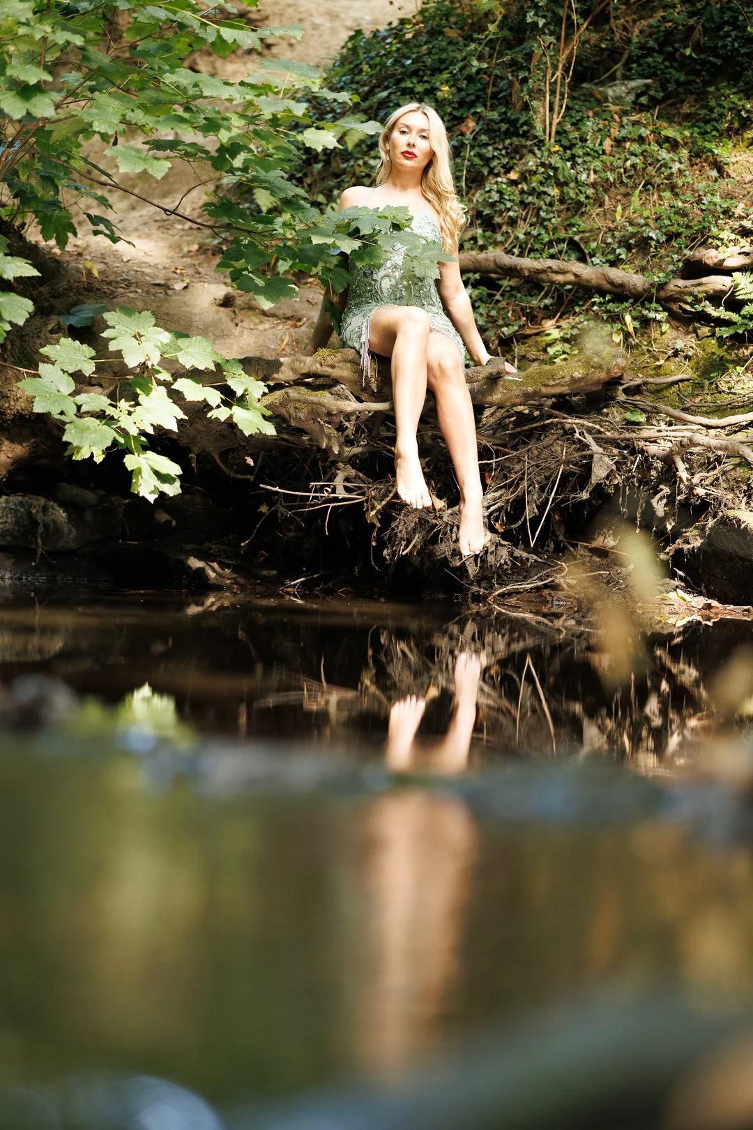 Blonde woman in a short green sequined dress sitting on a log by a forest stream, reflected in the water.