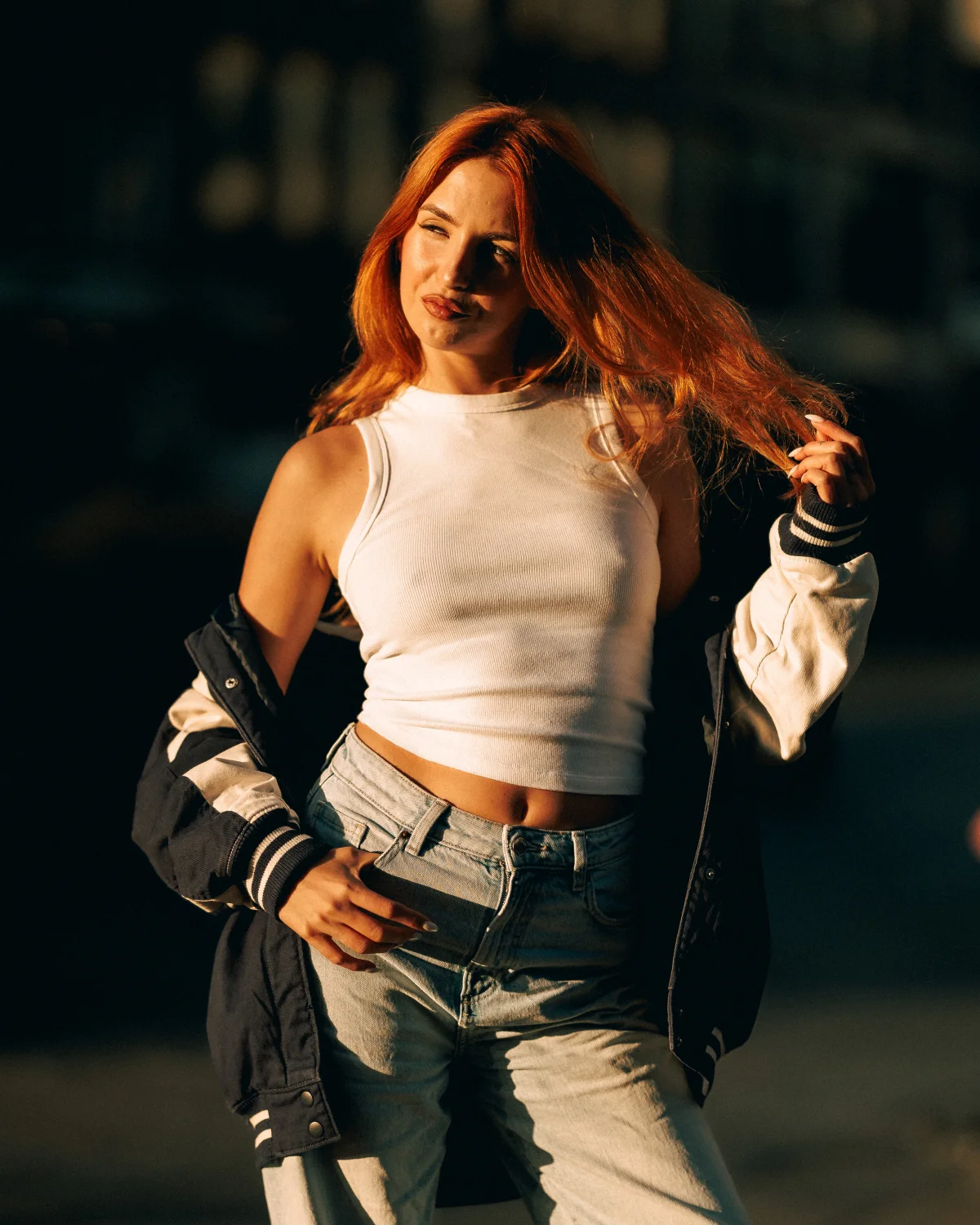 Woman with red hair in a white crop top and varsity jacket posing in golden hour sunlight.
