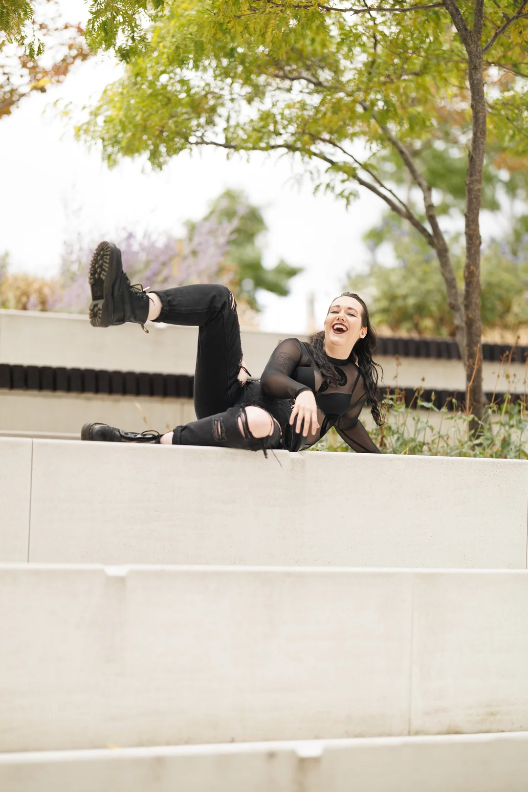 A woman in black attire laughing and posing dynamically on a concrete ledge outdoors.