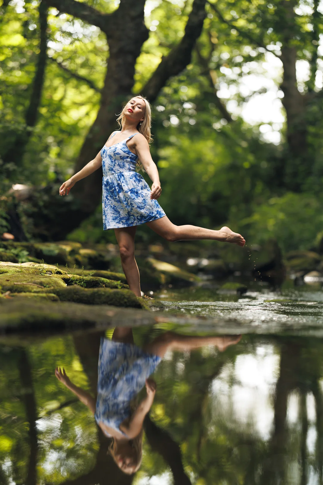 A woman in a blue floral dress poses gracefully by a forest stream, her reflection visible in the water.