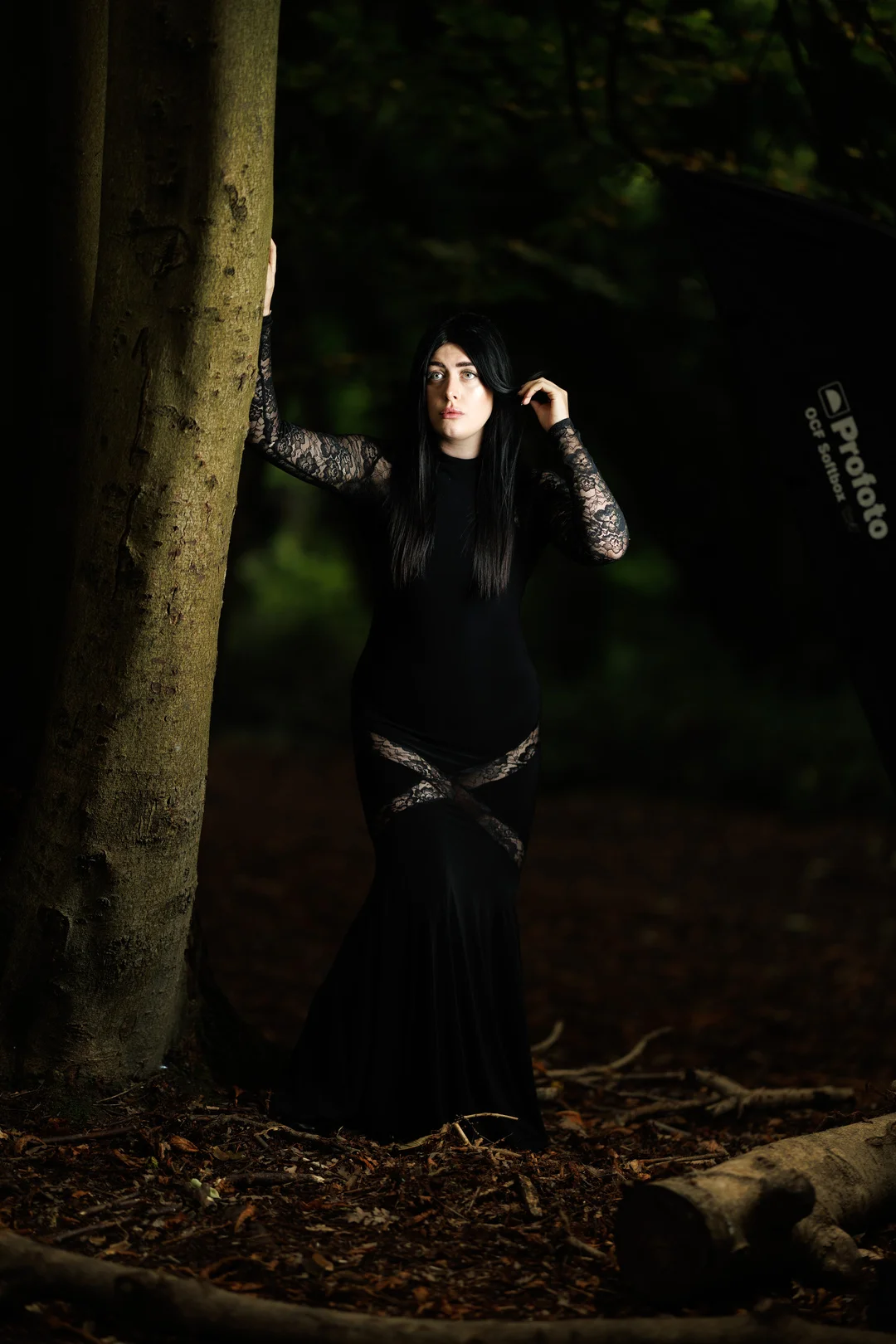 A woman with long black hair in a long black lace dress posing against a tree in a moody, dark forest.