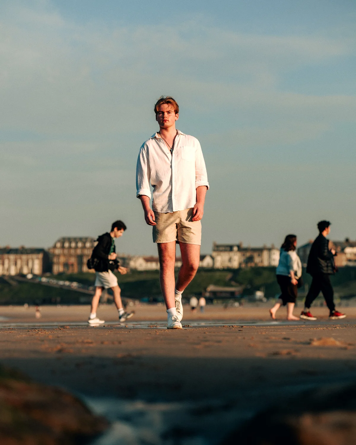 A young man in a white linen shirt and beige shorts walking on a beach during golden hour.