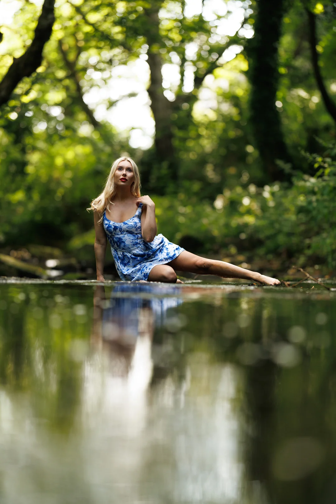 Blonde woman in a blue floral dress posing in a shallow forest stream with a reflection in the water.