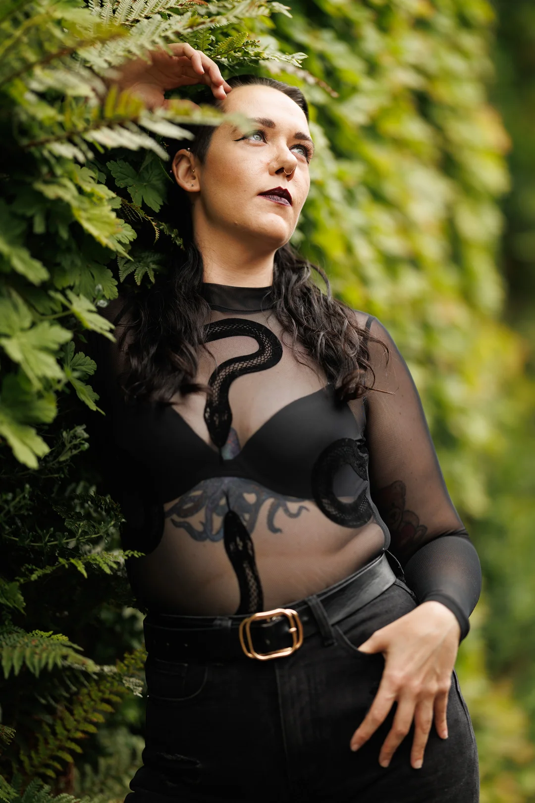 A person with dark hair and bold makeup posing against a lush green hedge, wearing a sheer black snake-print top.