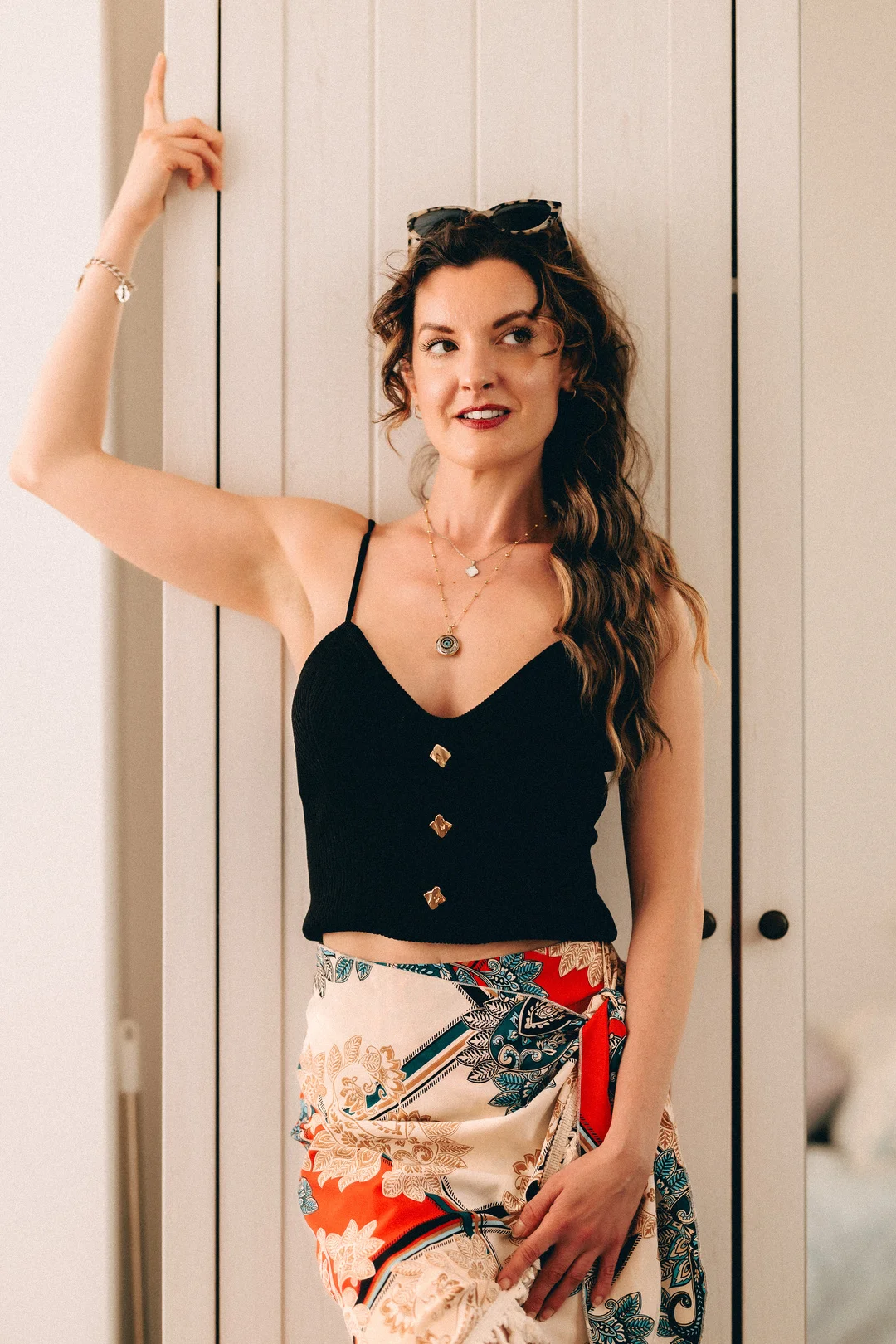 A woman with long wavy hair wearing a black top and a colorful patterned skirt, posing against a white paneled wall.
