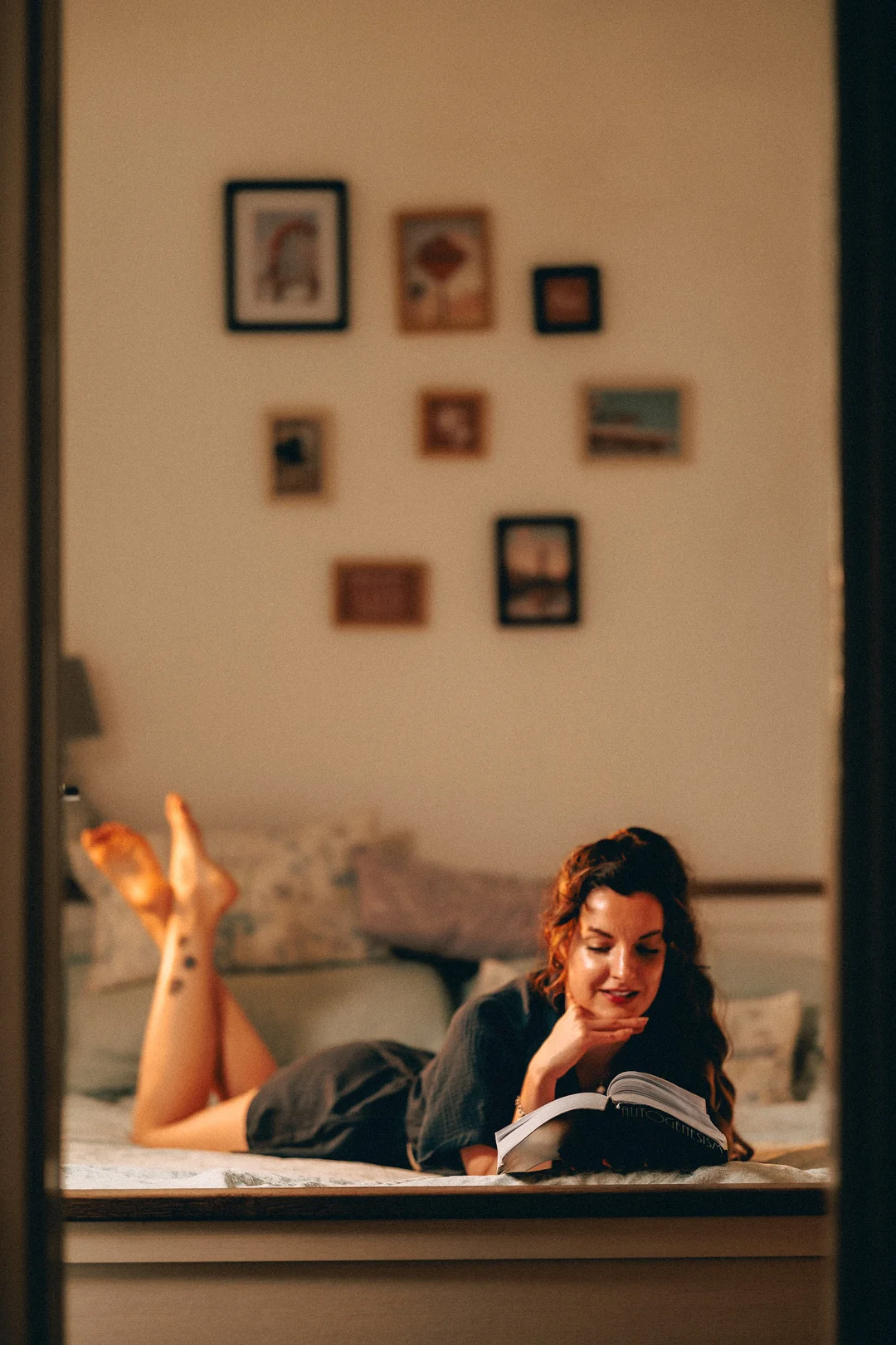 A woman lying on a bed reading a book in a cozy, warmly lit room with art on the wall.