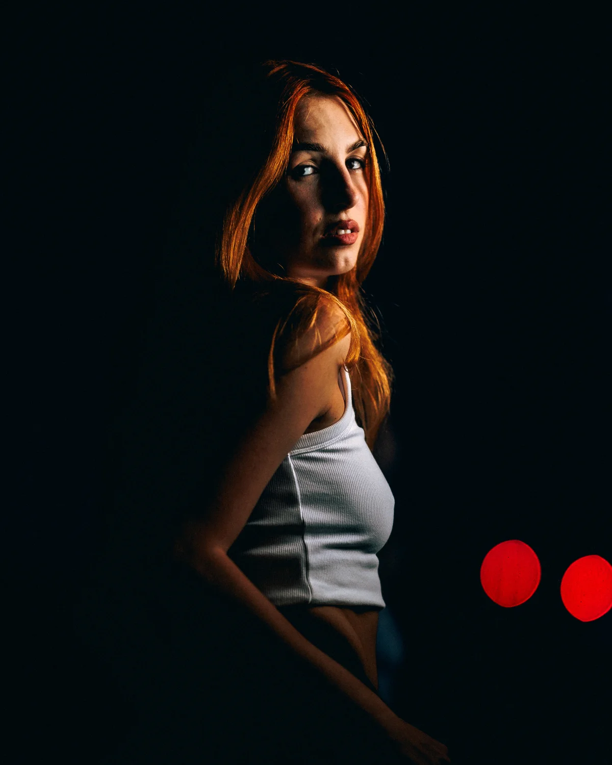Moody portrait of a red-haired woman in a white tank top against a dark background with red lights.