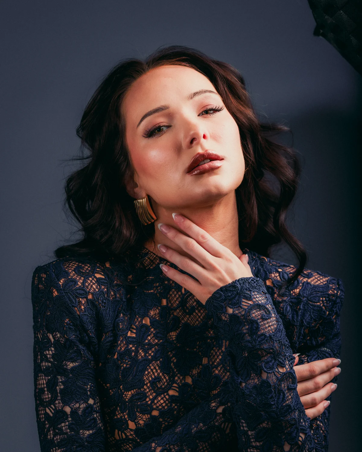 A woman with dark wavy hair and bold makeup posing with her hand on her neck, wearing a navy blue lace dress.