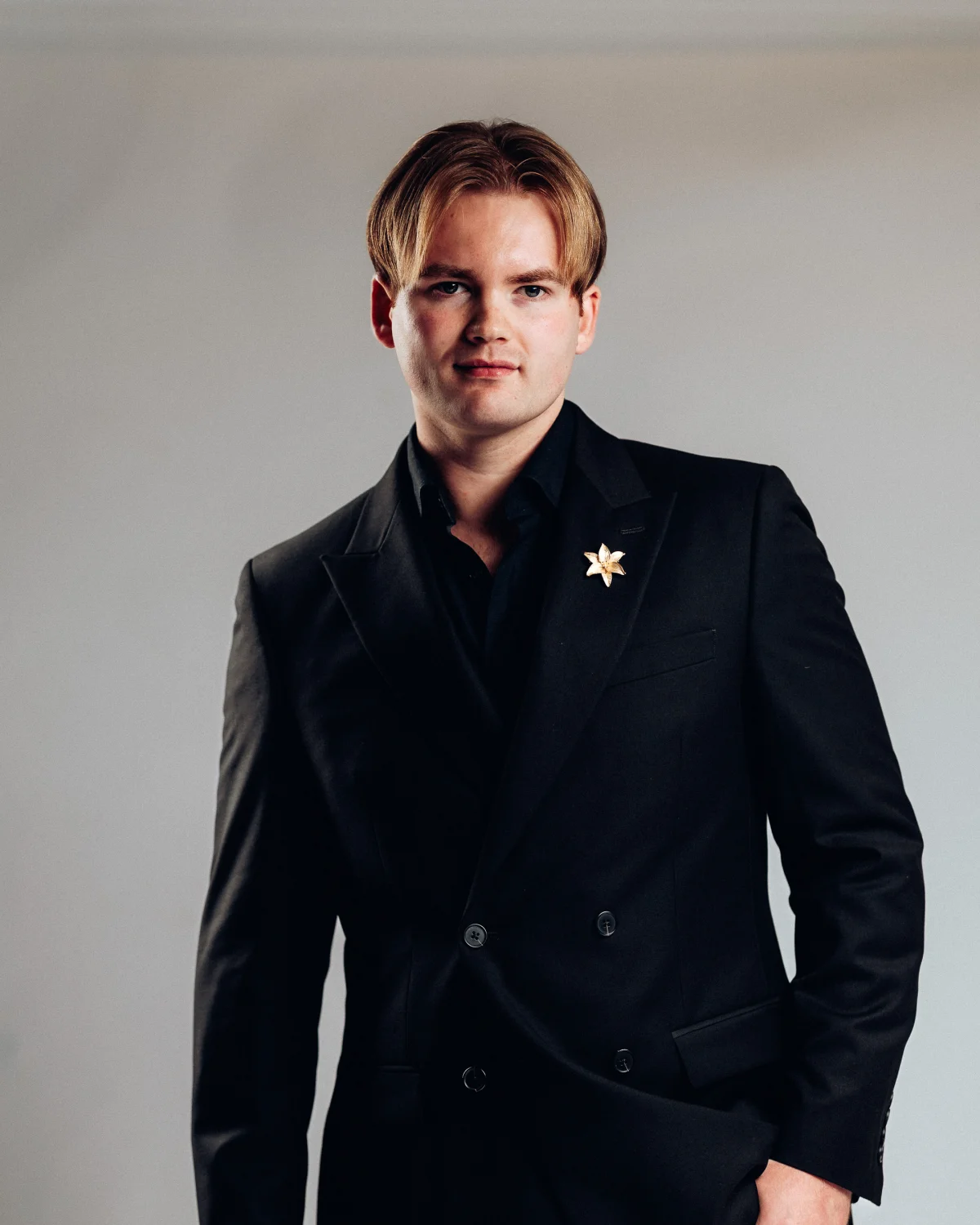 A professional portrait of a man with blond hair in a black double-breasted suit against a neutral grey background.