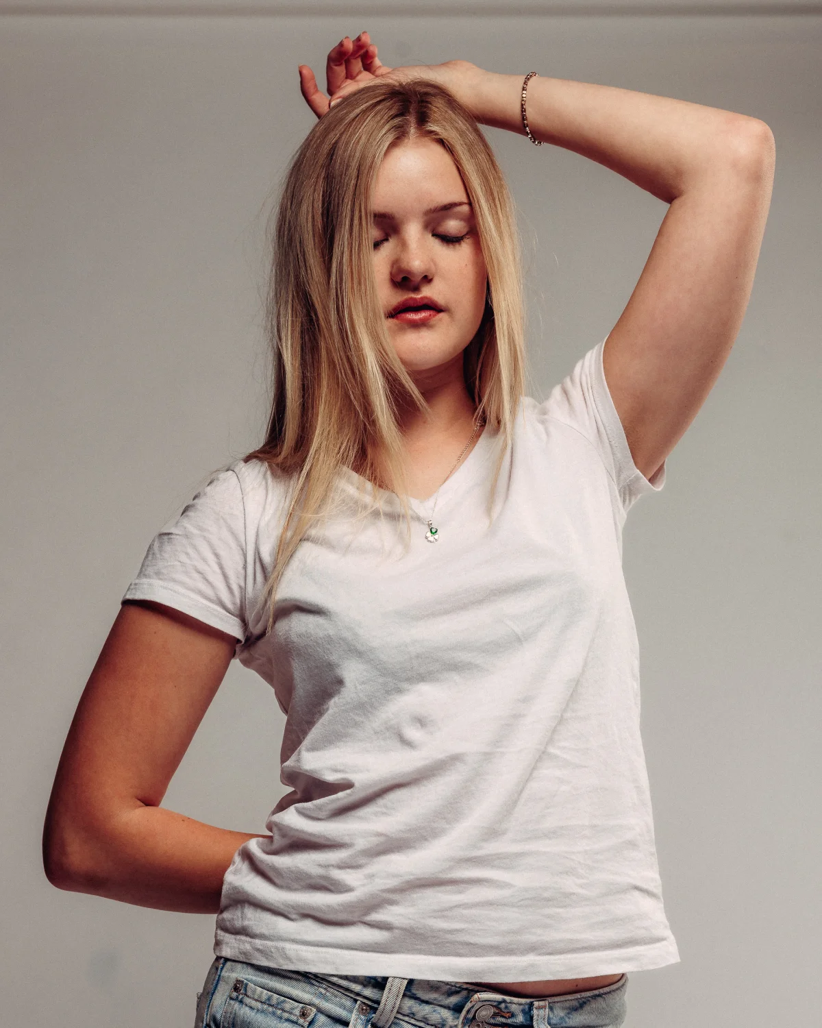 A young woman with blonde hair in a white t-shirt and jeans, posing with eyes closed against a neutral background.