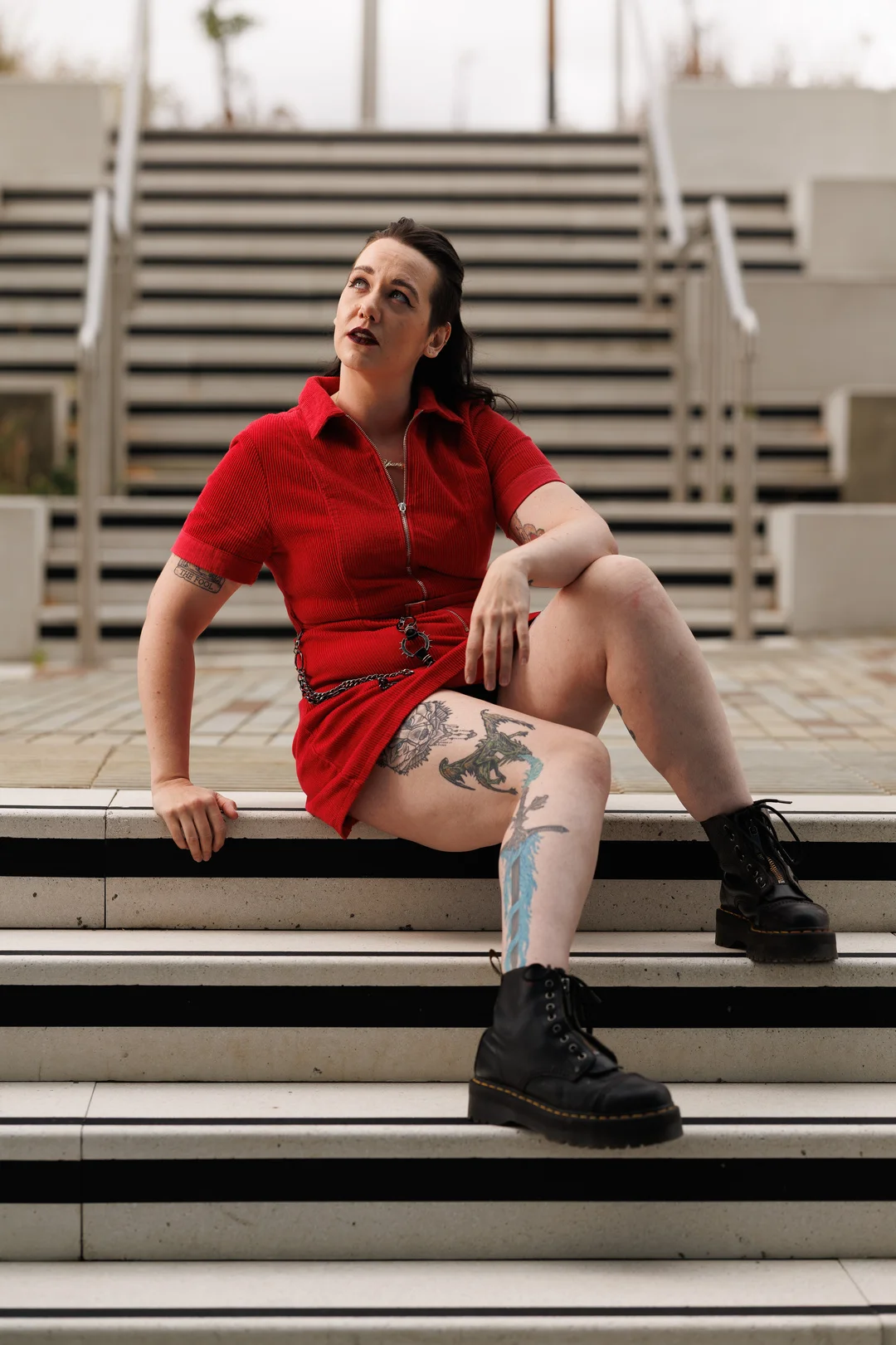 Woman with tattoos in a red dress and black boots sitting on outdoor concrete stairs.