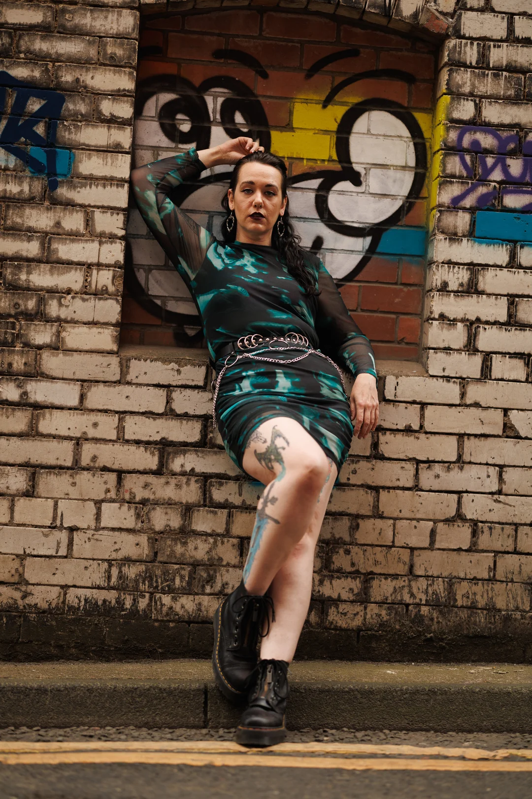 A person with tattoos in a dark green patterned dress and black boots posing against a graffiti brick wall.