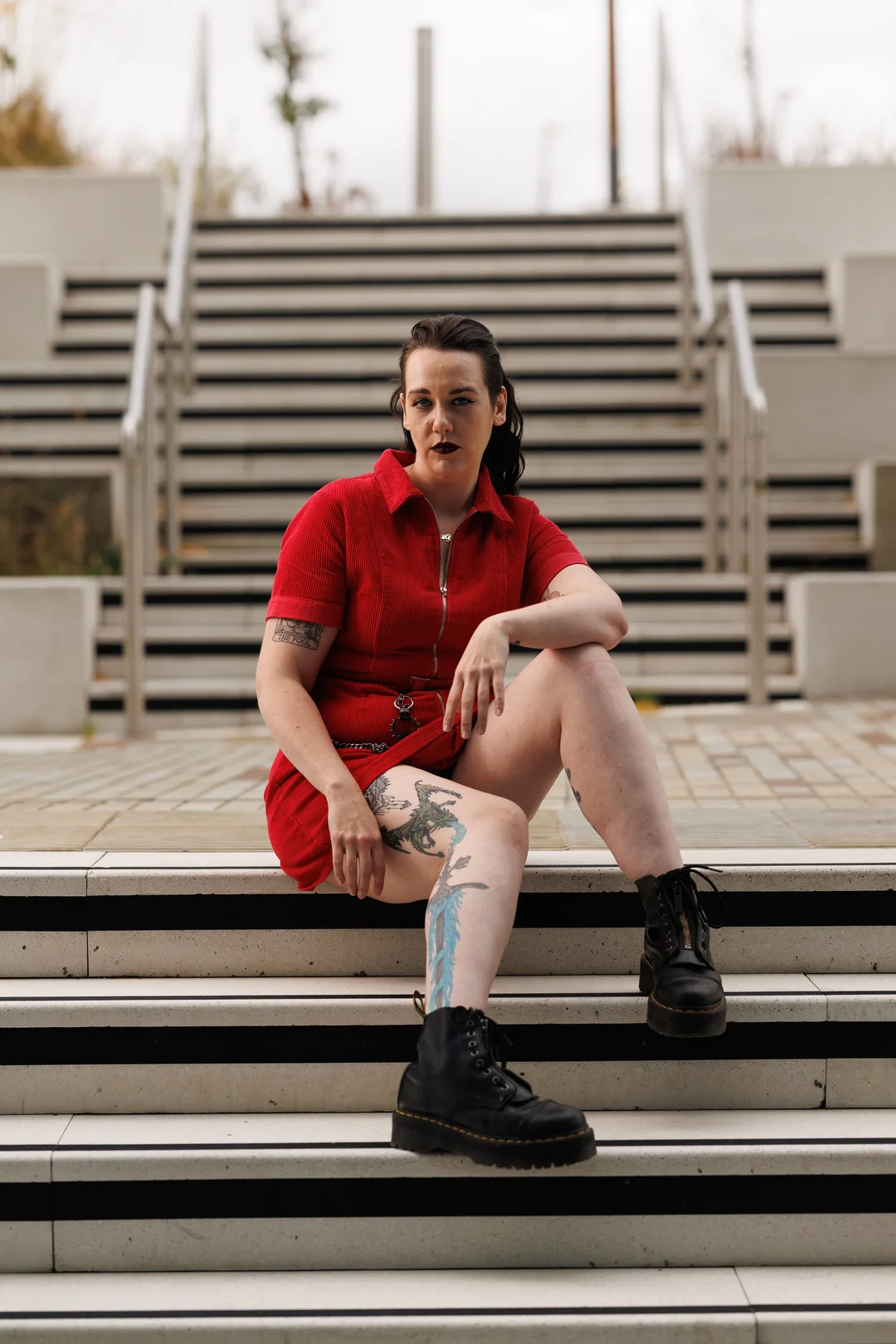 A person with tattoos in a red outfit and black boots sitting on outdoor concrete stairs.