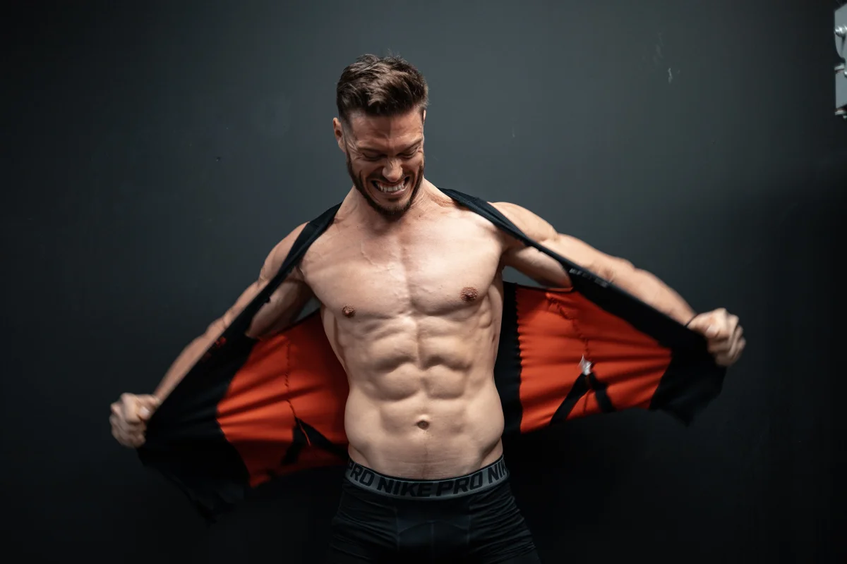 Muscular man pulling open a black and orange jacket to reveal his abs against a dark studio background.