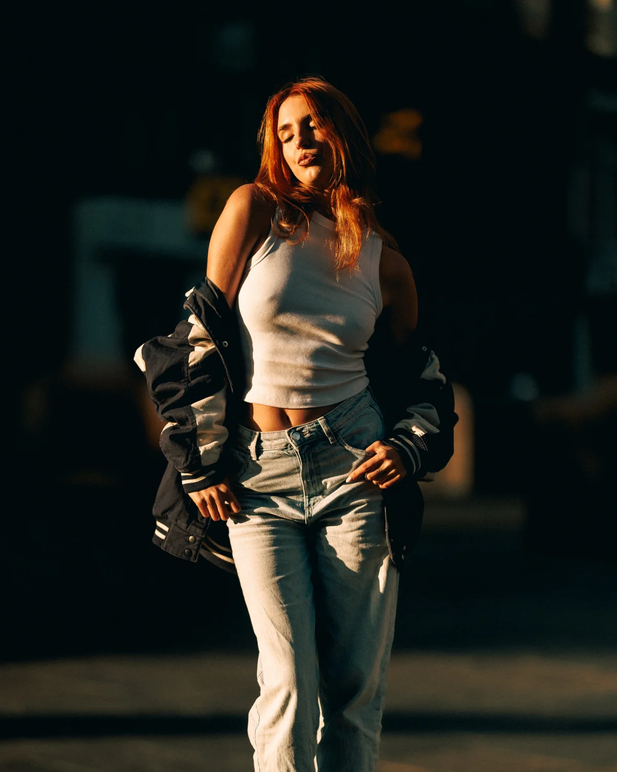 Young woman with red hair posing outdoors in a white tank top, jeans, and a varsity jacket draped off her shoulders.