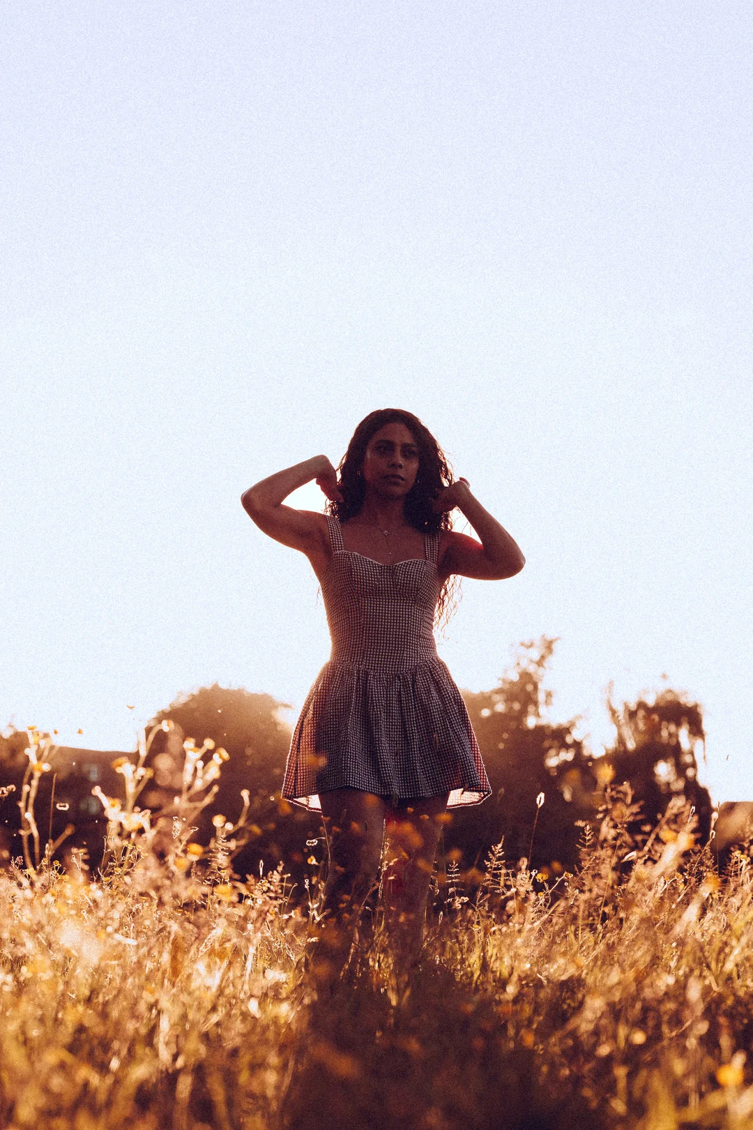 A woman in a red and white checkered dress posing in a golden field during sunset.