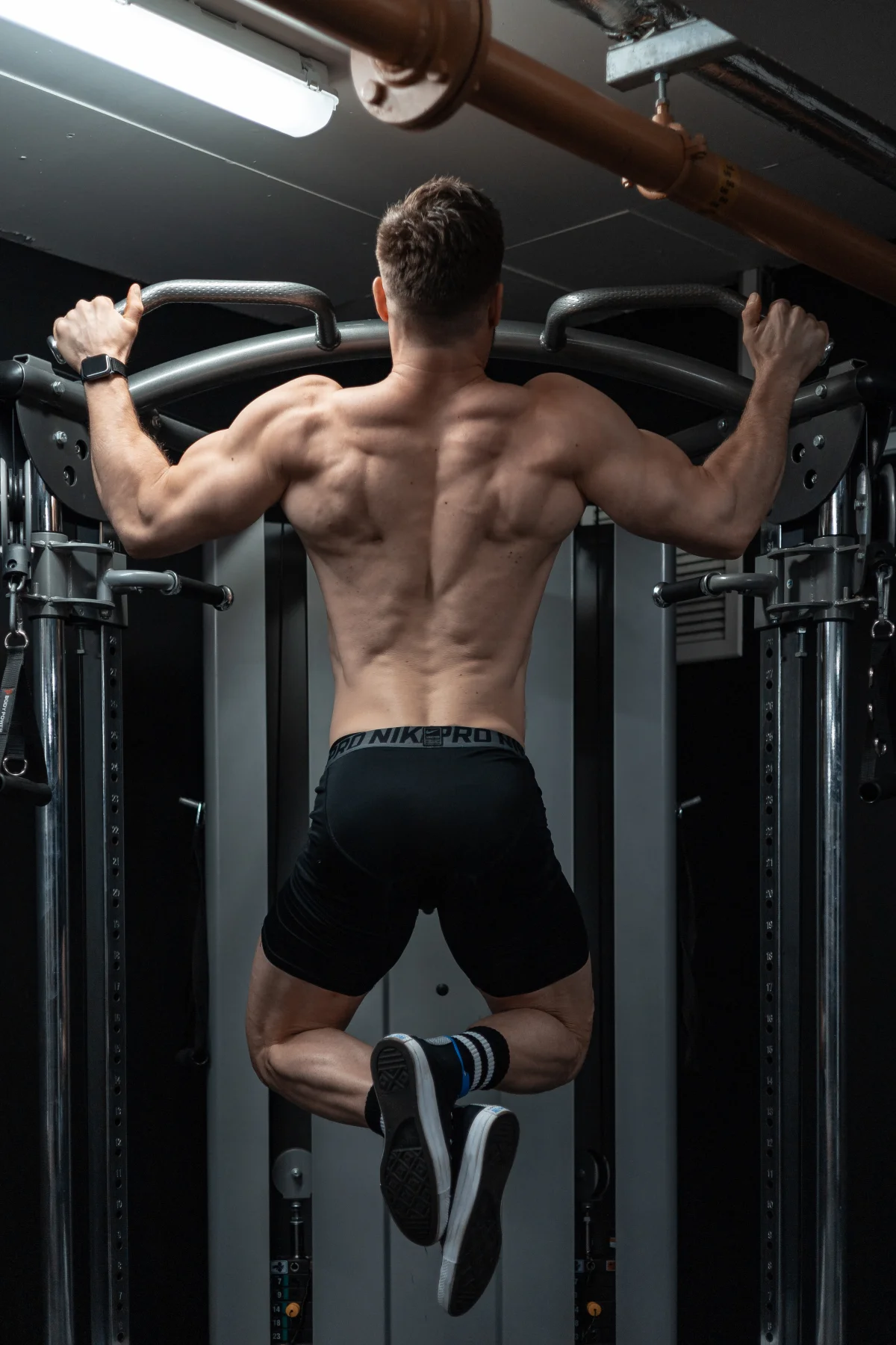 Muscular man performing a pull-up in a gym, viewed from behind to highlight back definition.