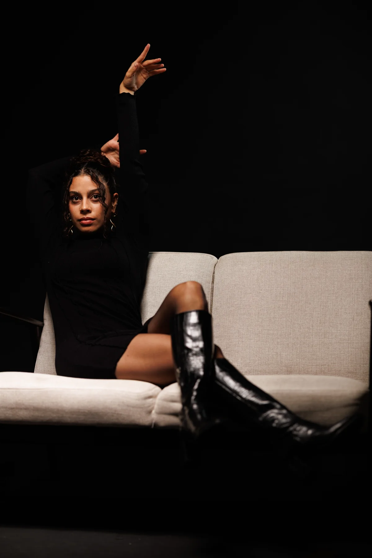 Woman in a black dress and glossy black boots posing on a white couch against a black background.