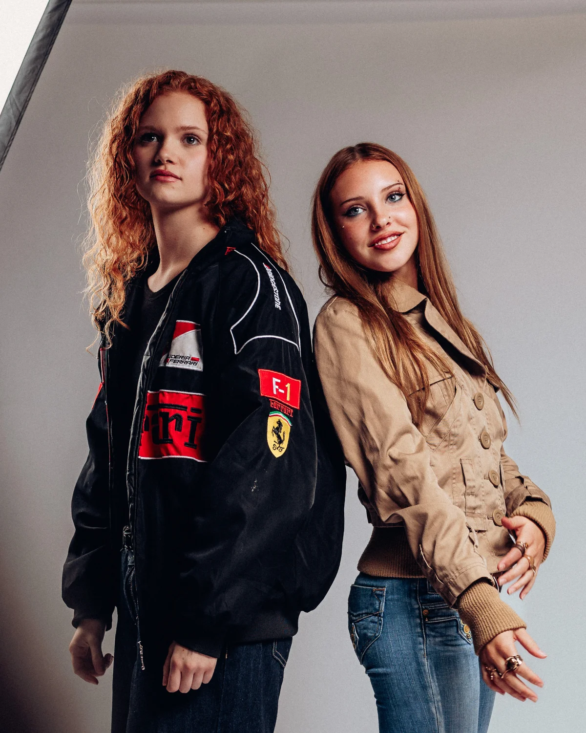 Two young women posing back-to-back in a studio, one in a black Ferrari racing jacket and one in a tan jacket.