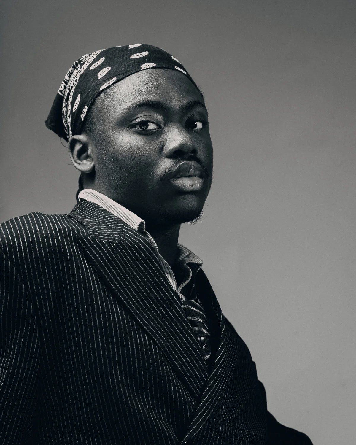 Black and white studio portrait of a Black man in a pinstripe suit and patterned bandana with a serious expression.