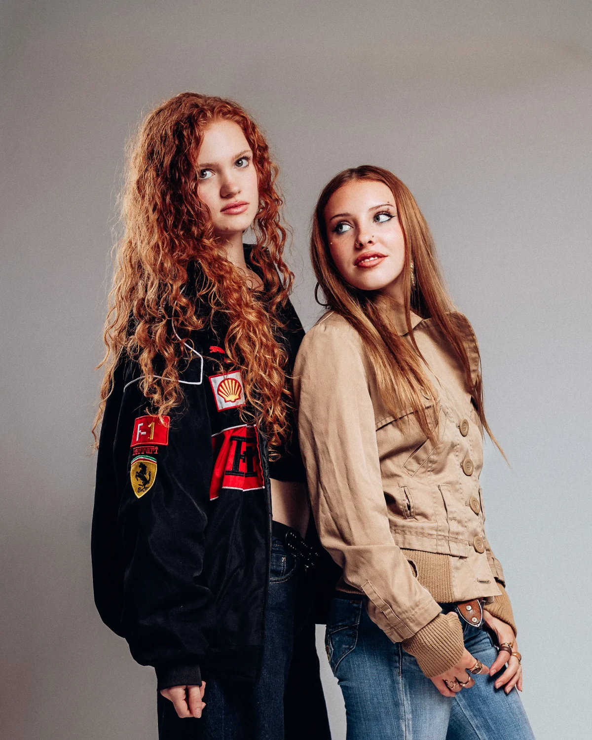 Two young women with red hair posing in a studio; one wears a black Ferrari racing jacket, the other a tan jacket.