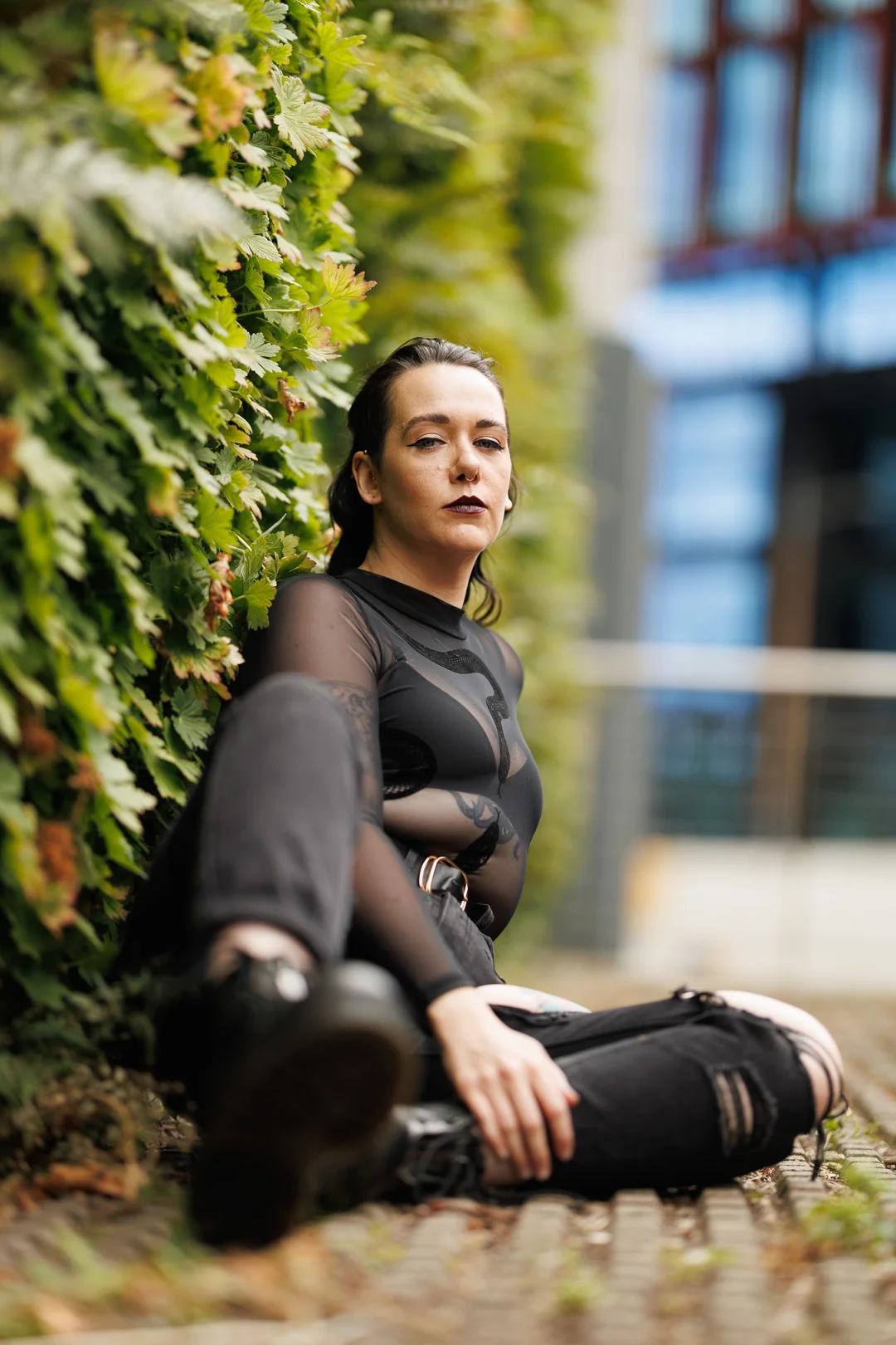 A person with dark makeup and a sheer black top sitting against a lush green hedge in an outdoor setting.