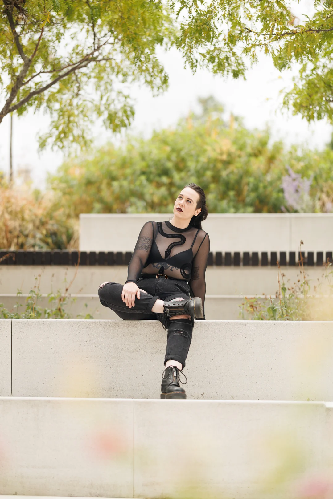 A person in alternative black fashion sitting on a concrete ledge in a garden setting.