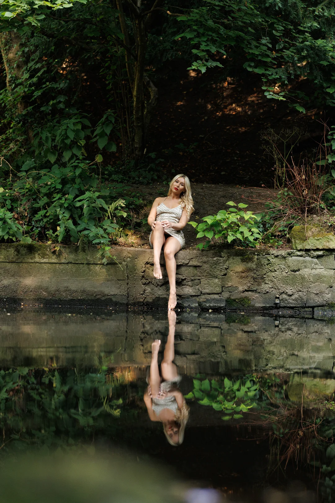 Woman in a shimmering gold dress sitting on a stone ledge by a pond with a clear reflection in the water.