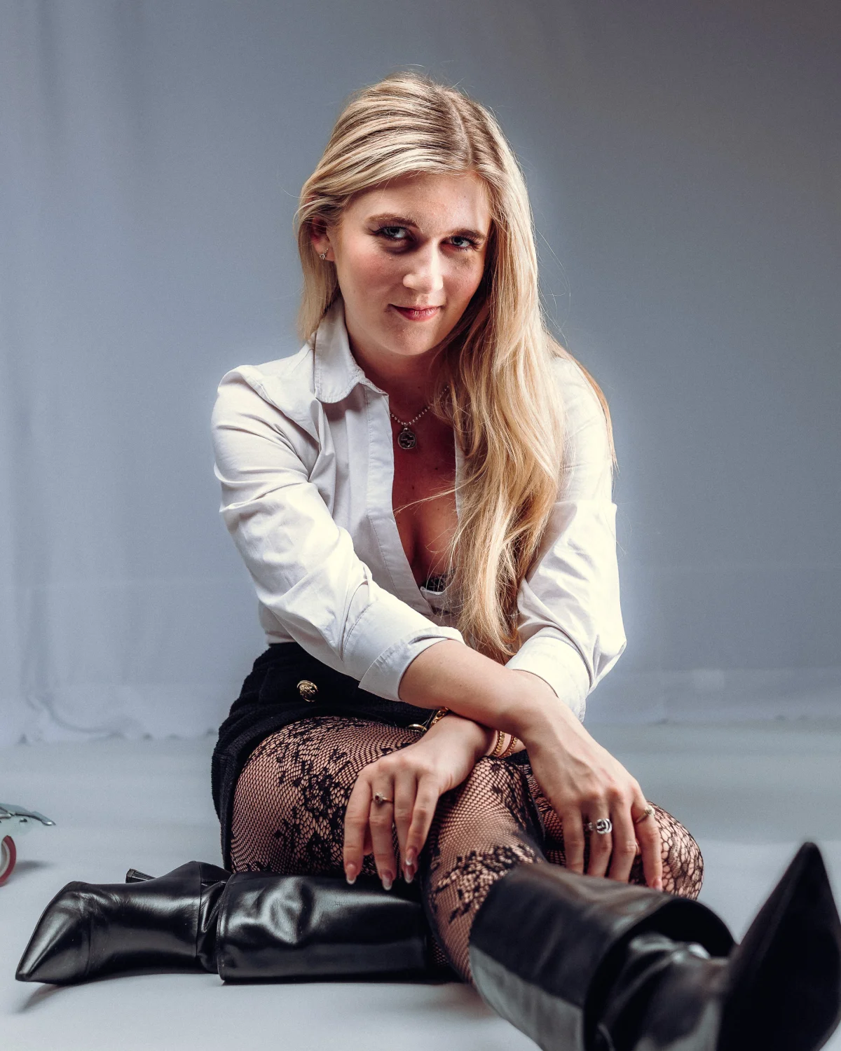 Blonde woman posing in a white shirt, black skirt, lace tights, and black boots against a grey studio background.