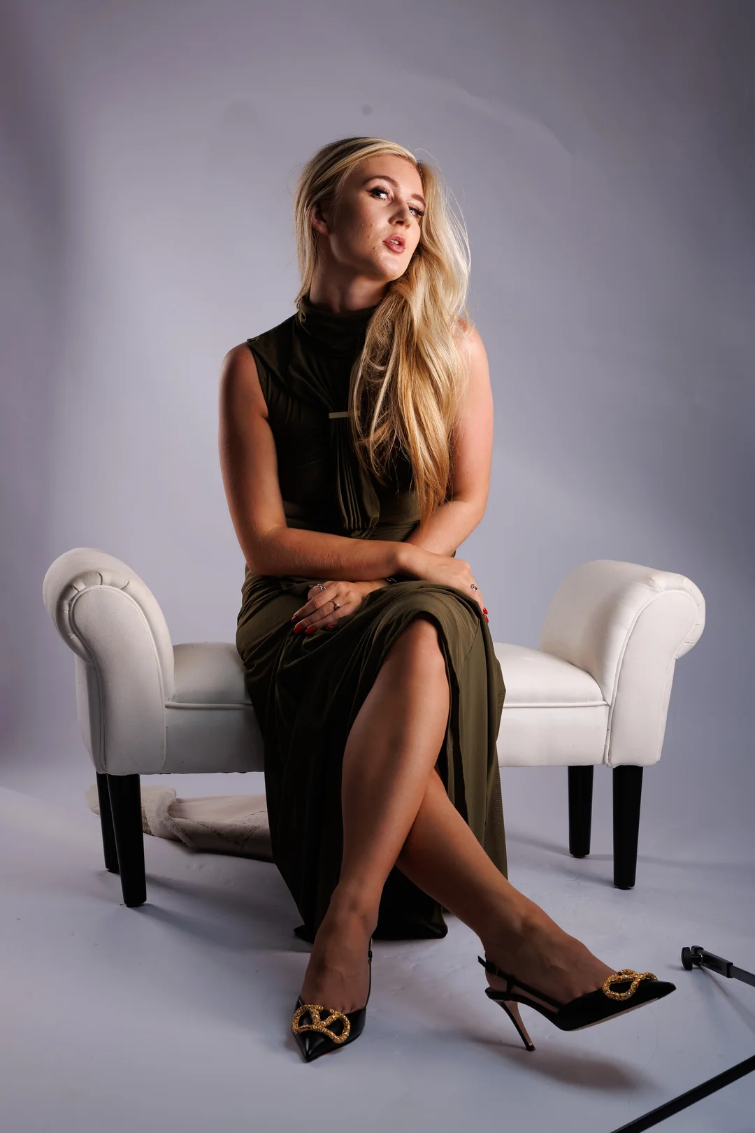 Blonde woman in an olive green dress posing on a white chaise lounge in a studio.