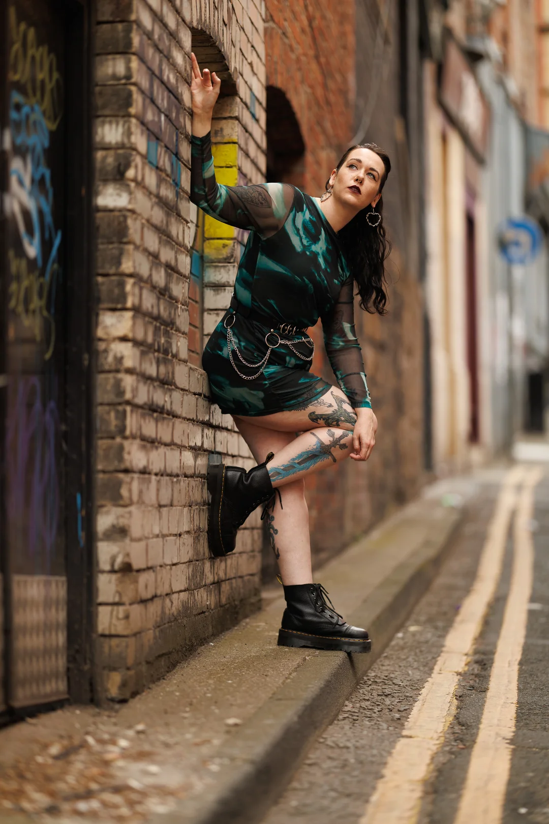 Woman with tattoos in a green patterned dress posing against a brick wall in an urban alley.