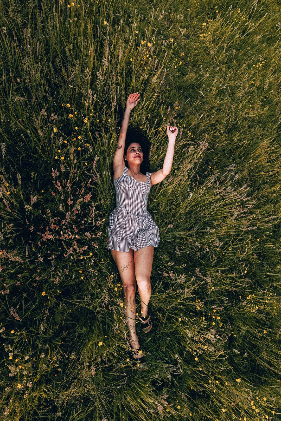 A woman in a gingham dress lying in a lush green field of grass, looking up with a serene expression.