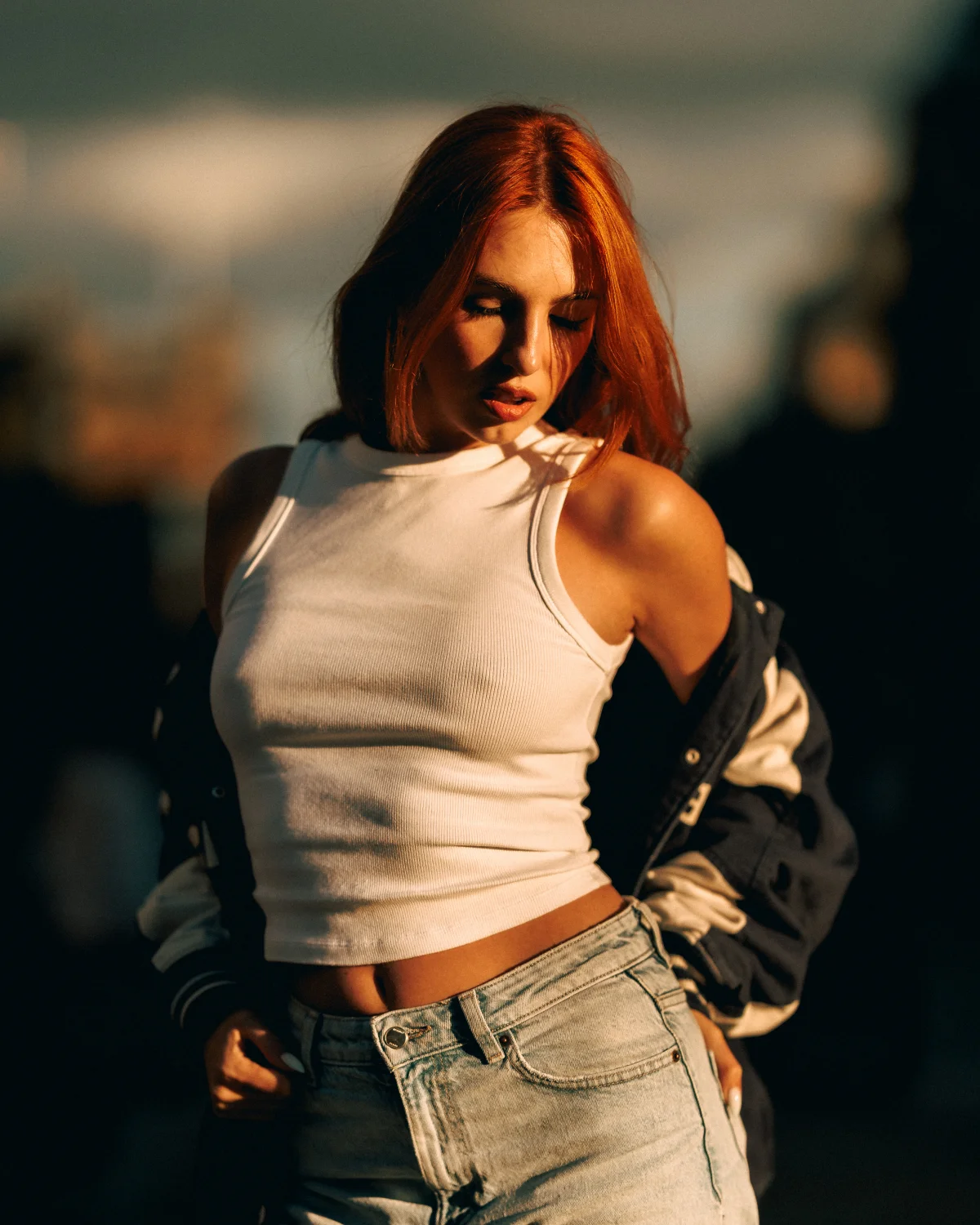 A woman with red hair in a white crop top and jeans posing outdoors in golden hour light.