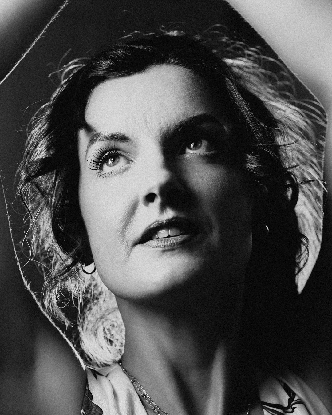 Black and white close-up portrait of a woman in a wide-brimmed hat looking upwards with a thoughtful expression.