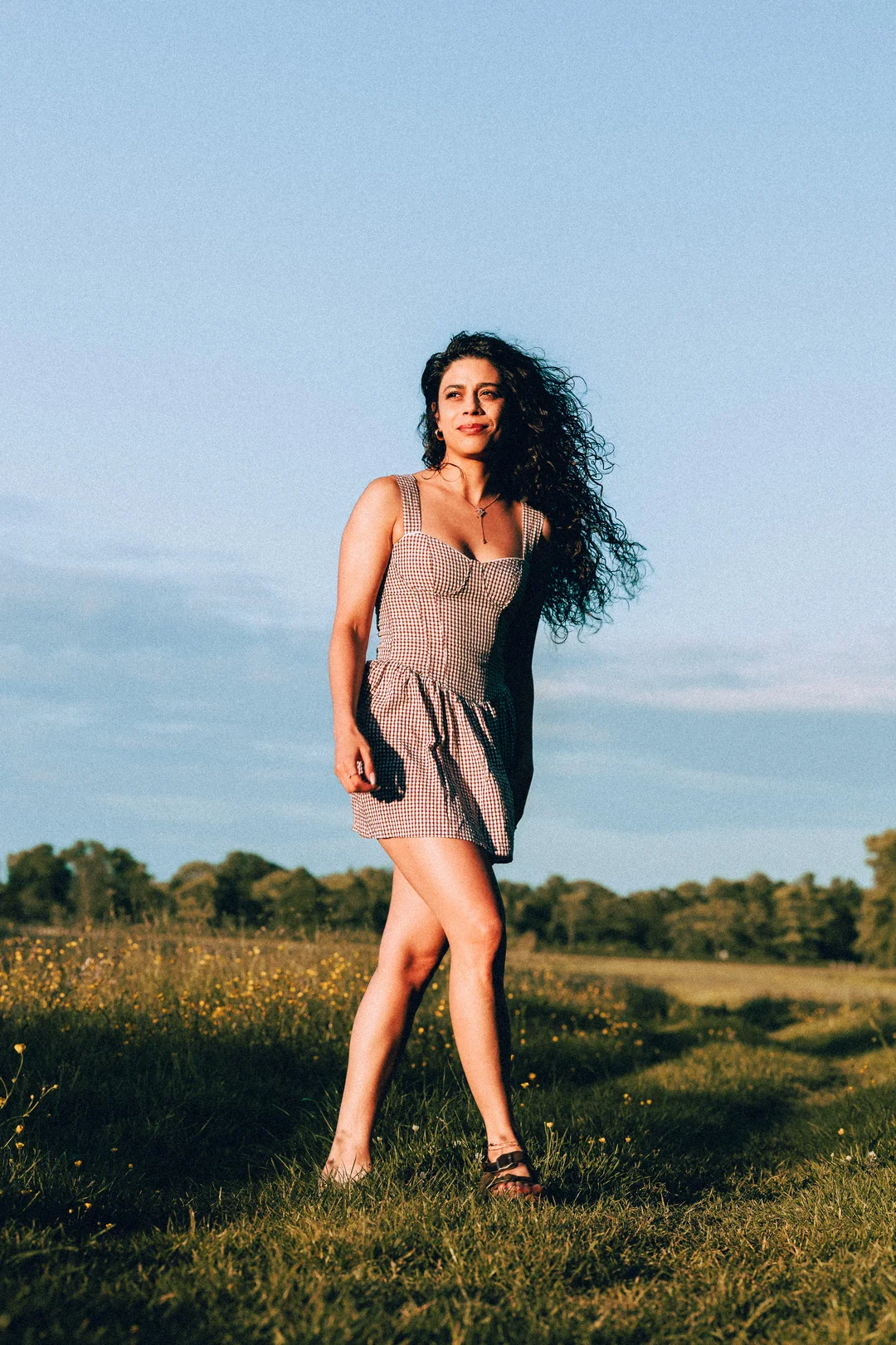 Woman with curly hair in a red and white gingham dress standing in a sunny grassy field under a blue sky.
