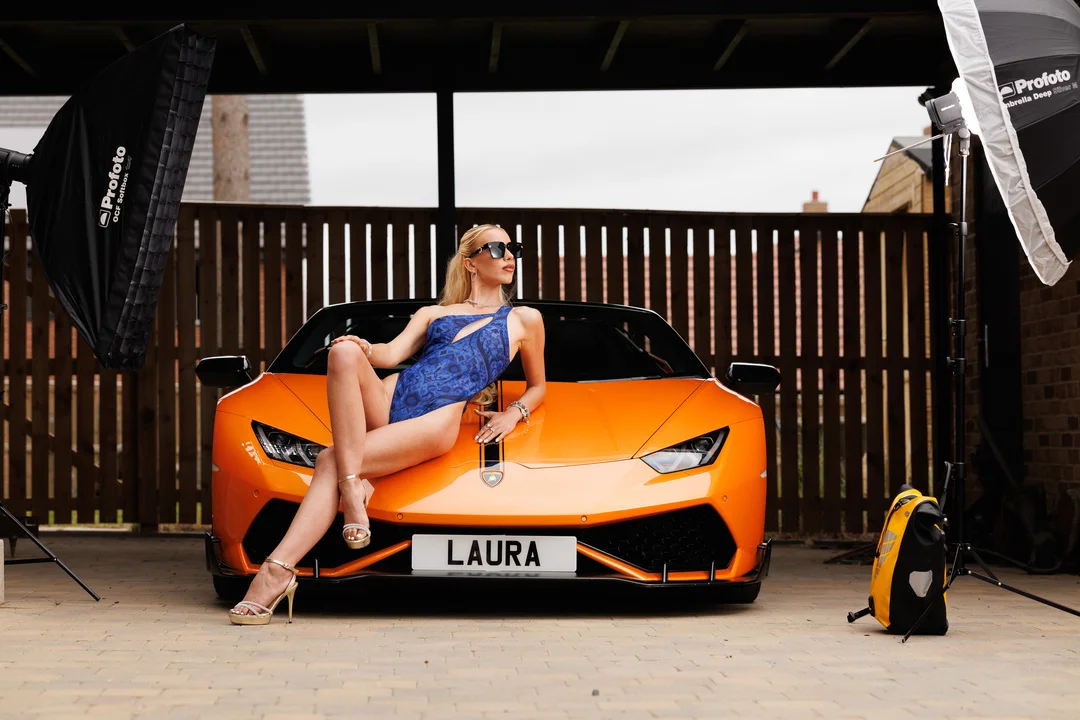 A woman in a blue swimsuit posing on the hood of an orange Lamborghini with studio lighting equipment visible.