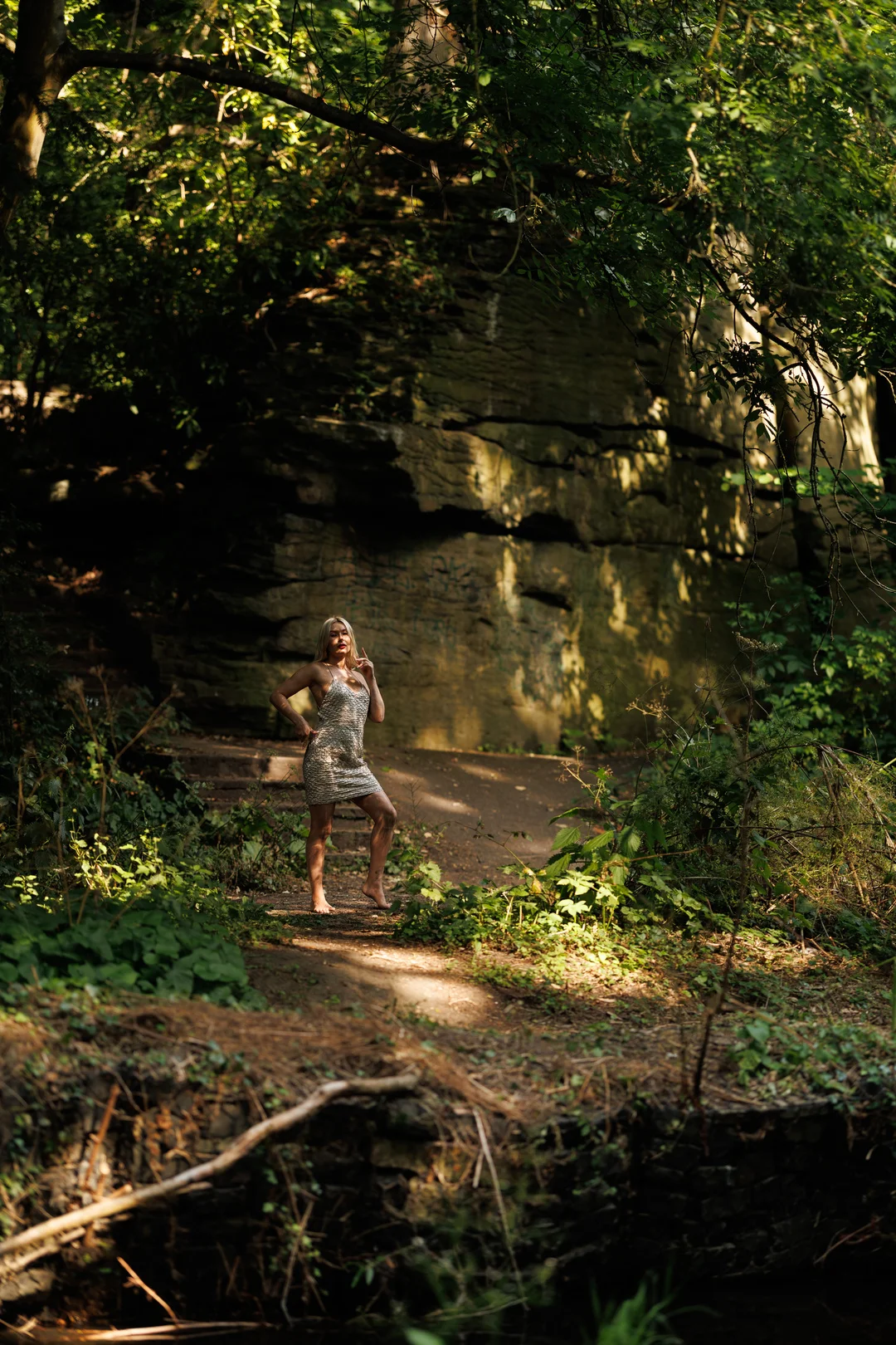 A woman in a short patterned dress posing in a wooded area in front of a large rock wall.
