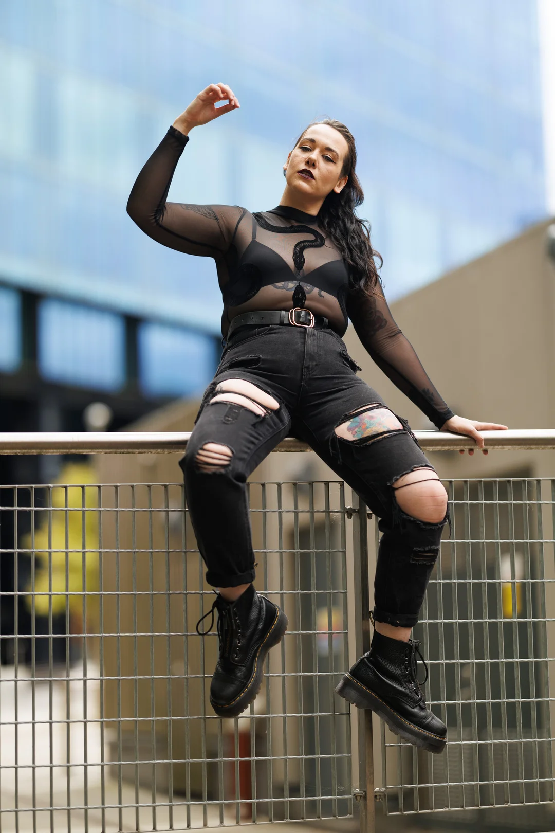 Woman in a black sheer top and ripped black jeans posing on a metal railing in an urban setting.
