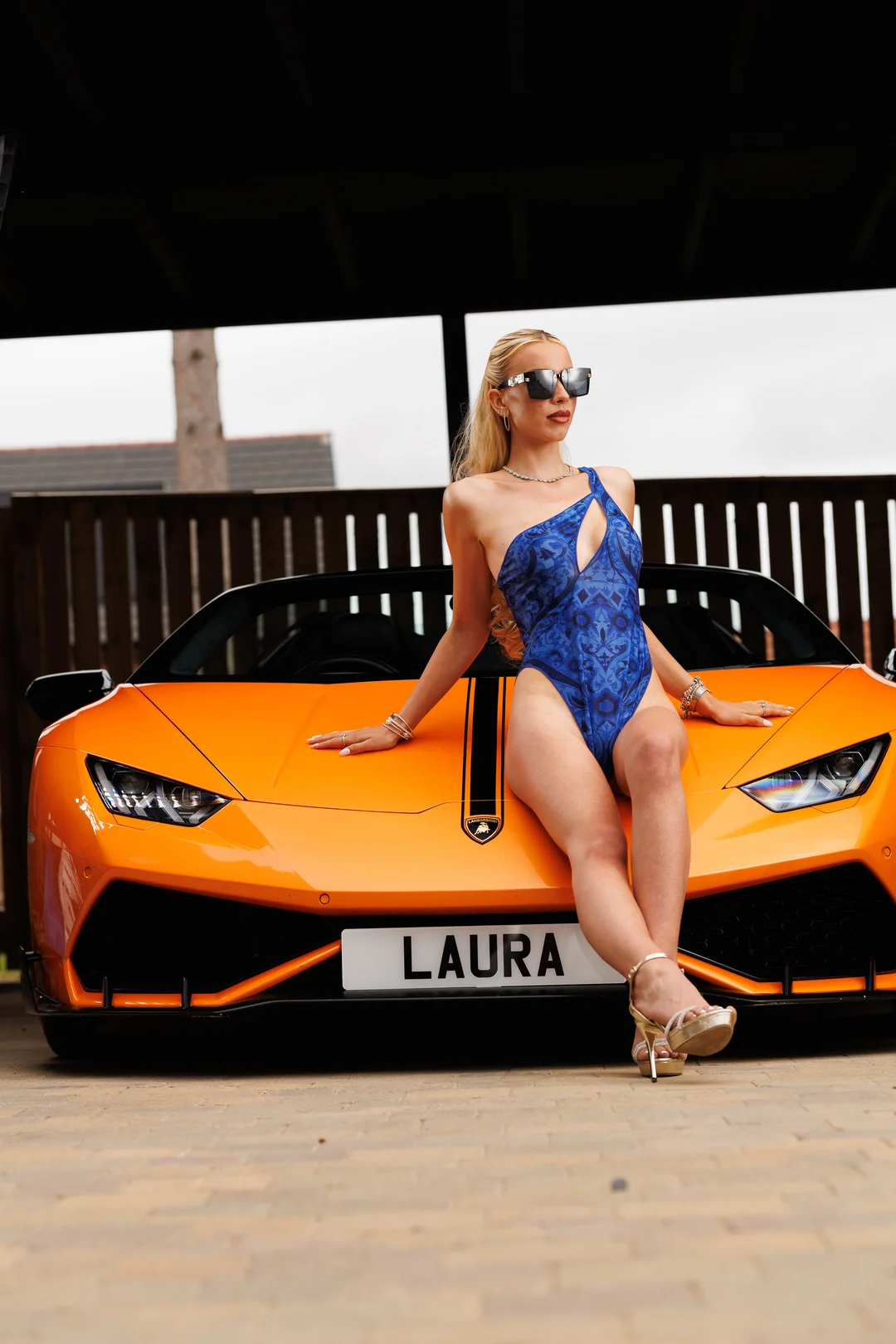 A blonde woman in a blue one-piece swimsuit and sunglasses posing on the hood of an orange Lamborghini.