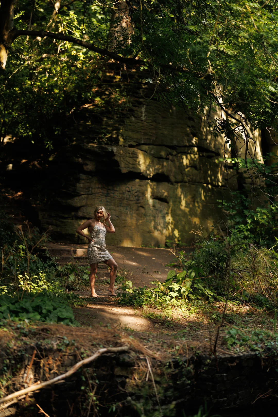 A woman in a short, shimmering dress posing in a sun-dappled forest in front of a large rock wall.