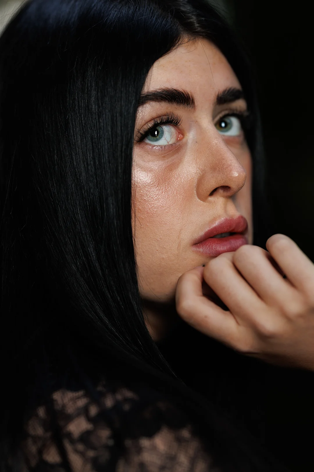 Close-up portrait of a woman with long black hair and blue eyes, looking thoughtfully with her hand near her lips.