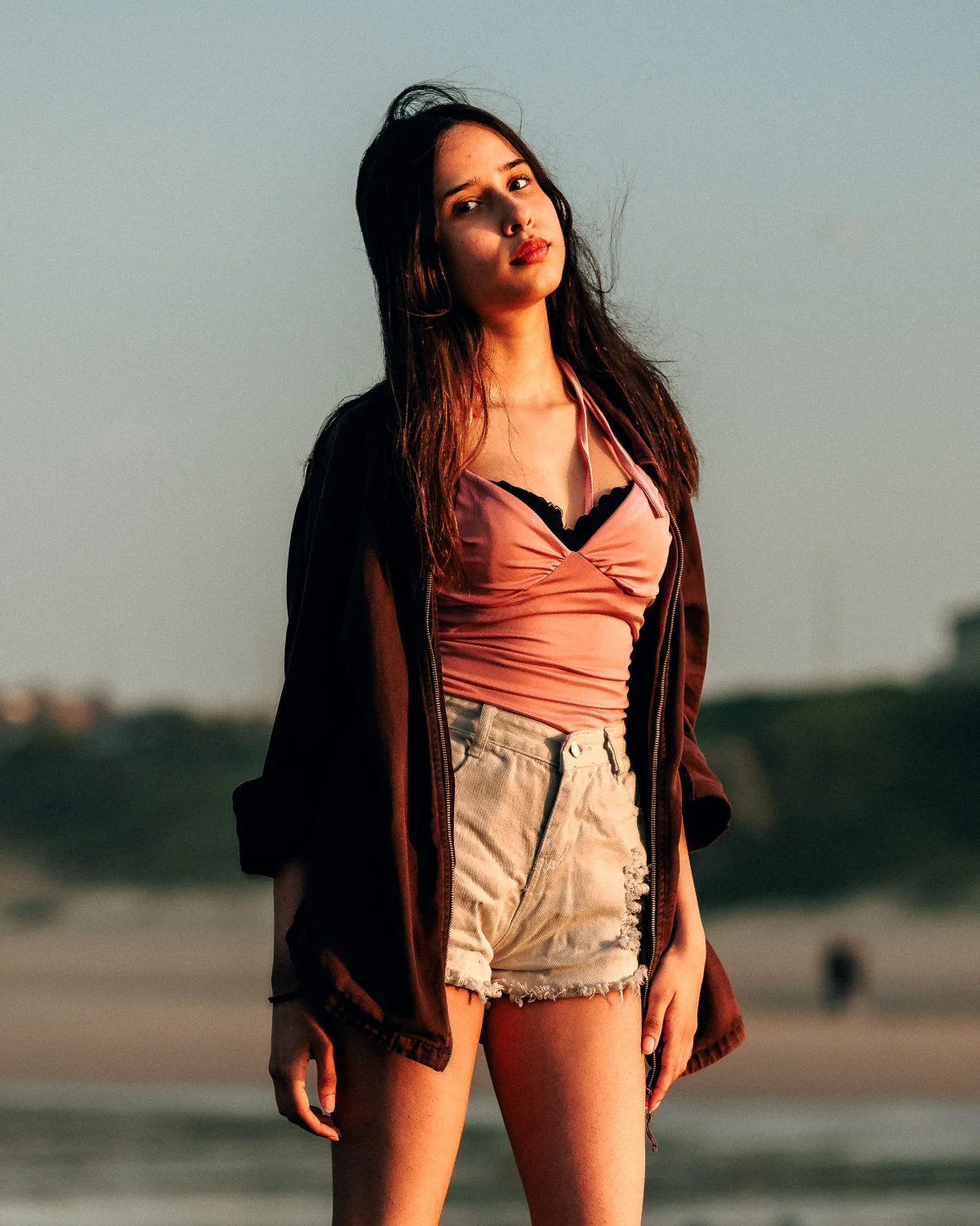 Young woman posing on a beach at sunset, wearing a pink top, white shorts, and a dark jacket.