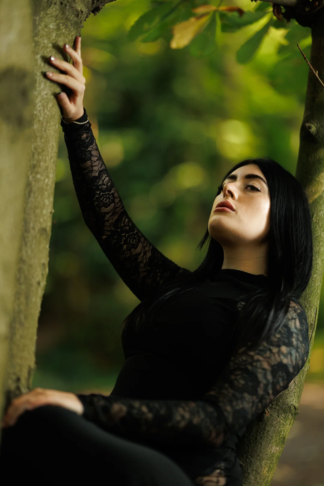 Woman with long black hair in a black lace dress leaning against a tree in a green forest.