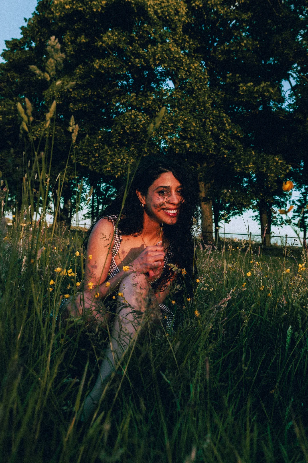 Smiling woman with curly hair sitting in a grassy field with yellow wildflowers and trees in the background.